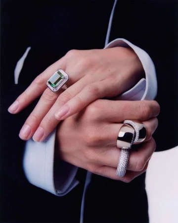 Close-up of an adult female's crossed hands showcasing rings with emerald-cut green gemstones and diamond accents set against a black suit.