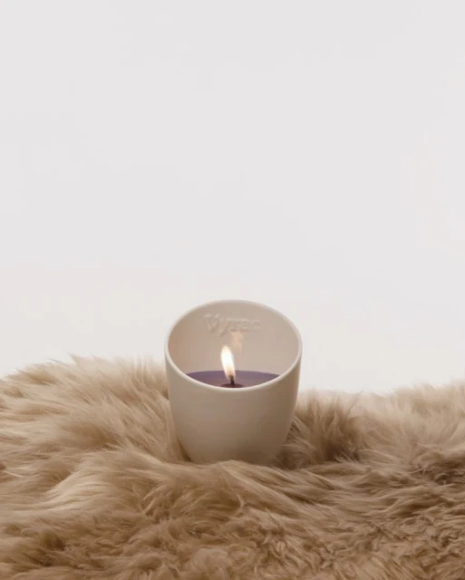 Lit candle in a white ceramic holder sits on a beige faux fur surface, creating a cozy and warm ambience, ideal for home decor.