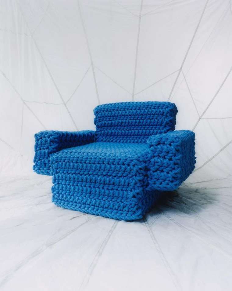 Blue miniature crocheted chair set against a white abstract backdrop. Studio lighting highlights the textured surface of the chair.