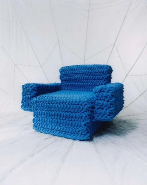 Blue miniature crocheted chair set against a white abstract backdrop. Studio lighting highlights the textured surface of the chair.