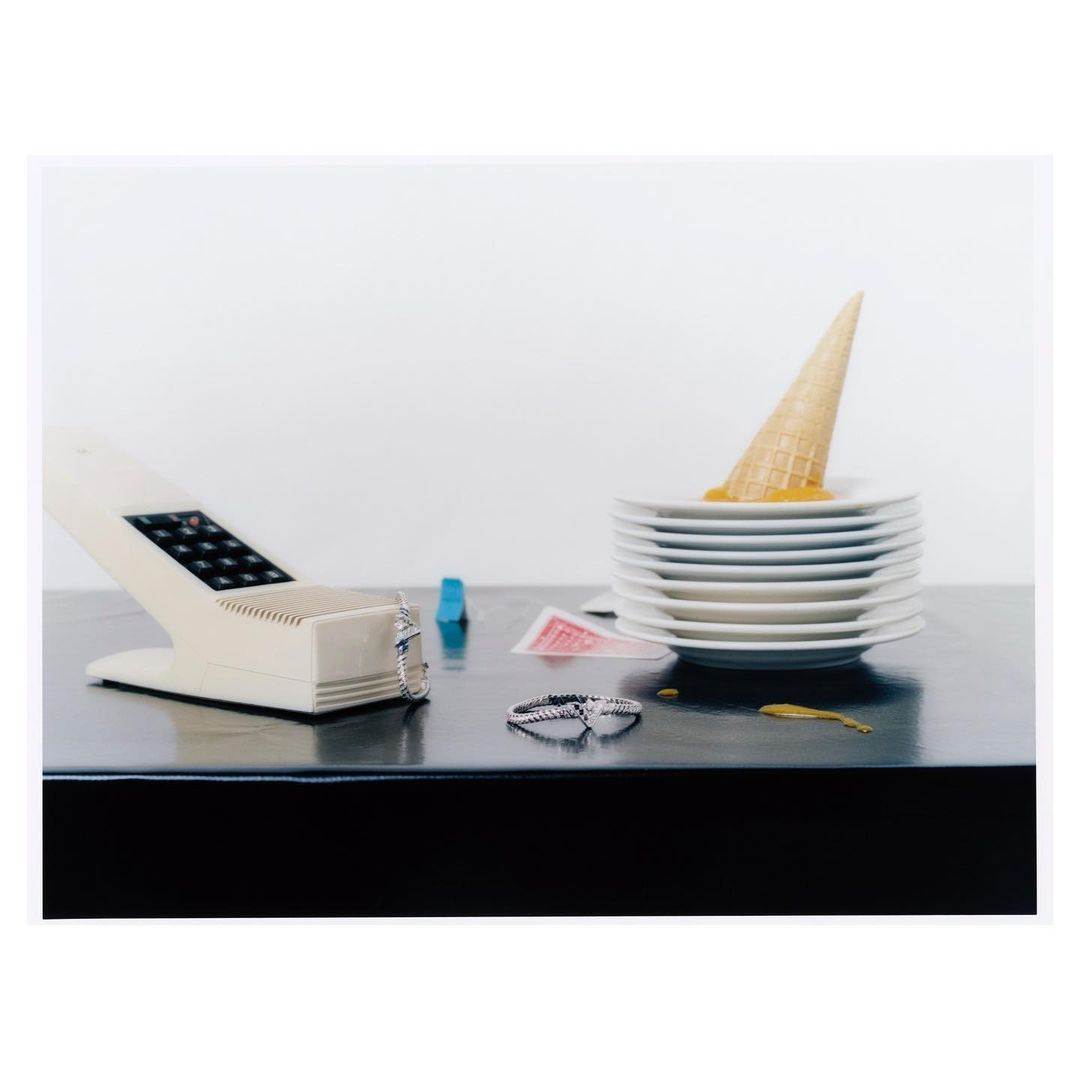 Still life of melted ice cream cone on stack of plates, vintage phone, rings, and playing cards on a black glossy surface against a white background.