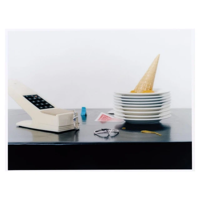 Still life of melted ice cream cone on stack of plates, vintage phone, rings, and playing cards on a black glossy surface against a white background.