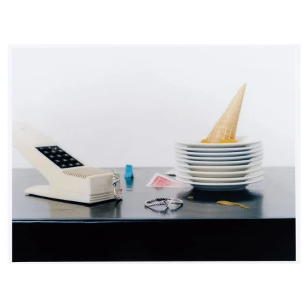 Still life of melted ice cream cone on stack of plates, vintage phone, rings, and playing cards on a black glossy surface against a white background.