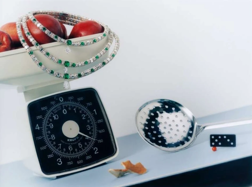 Still life studio shot of a diamond and emerald necklace draped over red plums in a container on a vintage kitchen scale.