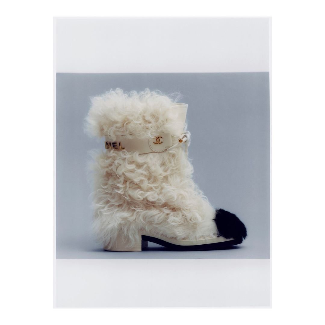 Fashion shot of a single ankle boot with off-white shearling, a black toe, and a decorative ankle strap against a soft grey backdrop.