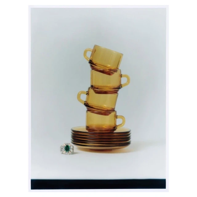 Stack of amber-colored glass teacups and saucers on a white surface with a diamond and emerald ring in the foreground.