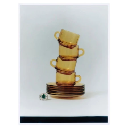 Stack of amber-colored glass teacups and saucers on a white surface with a diamond and emerald ring in the foreground.