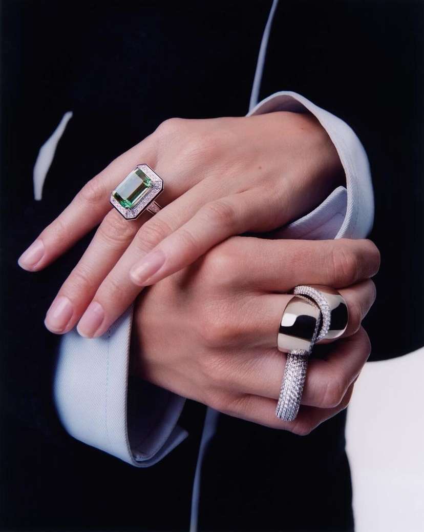 Close-up of hands with luxury rings. One hand features a large emerald-cut green gemstone ring, and the other has a modern silver band.