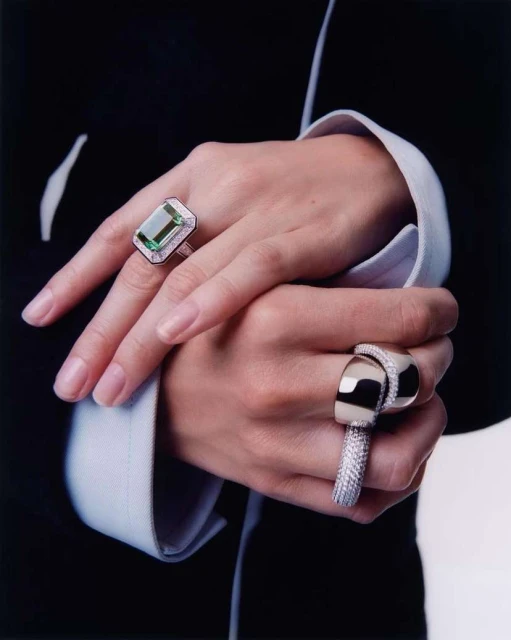 Close-up of hands with luxury rings. One hand features a large emerald-cut green gemstone ring, and the other has a modern silver band.