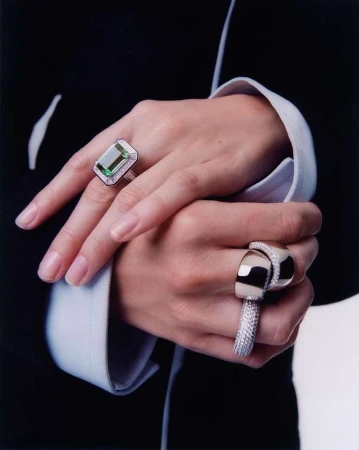 Close-up of hands with luxury rings. One hand features a large emerald-cut green gemstone ring, and the other has a modern silver band.
