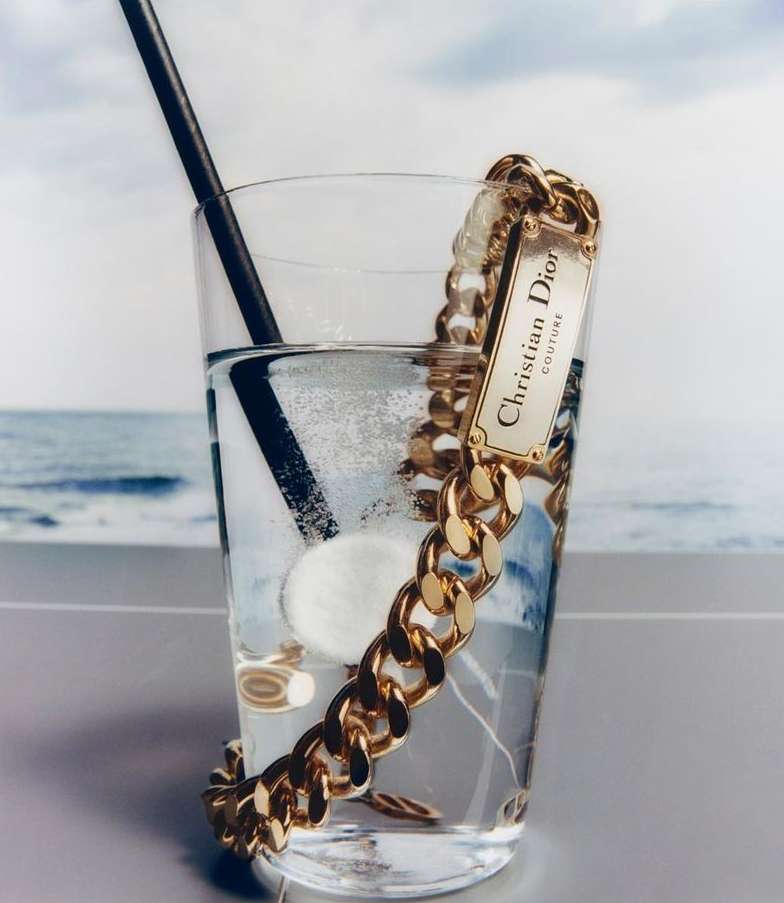Gold chain necklace with a Christian Dior logo plate drapes across a glass of clear liquid with a black straw, against a beach backdrop.