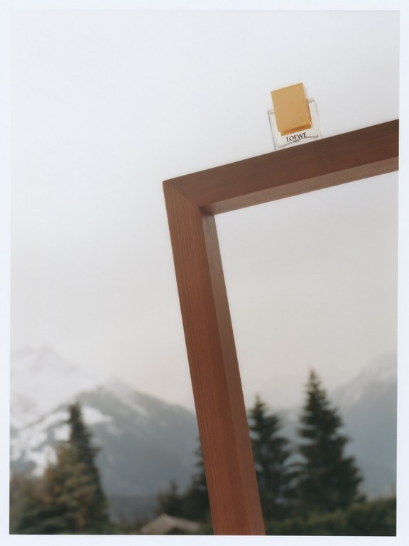 Loewe perfume bottle displayed on the edge of a picture frame, set against a blurred backdrop of mountains and trees.