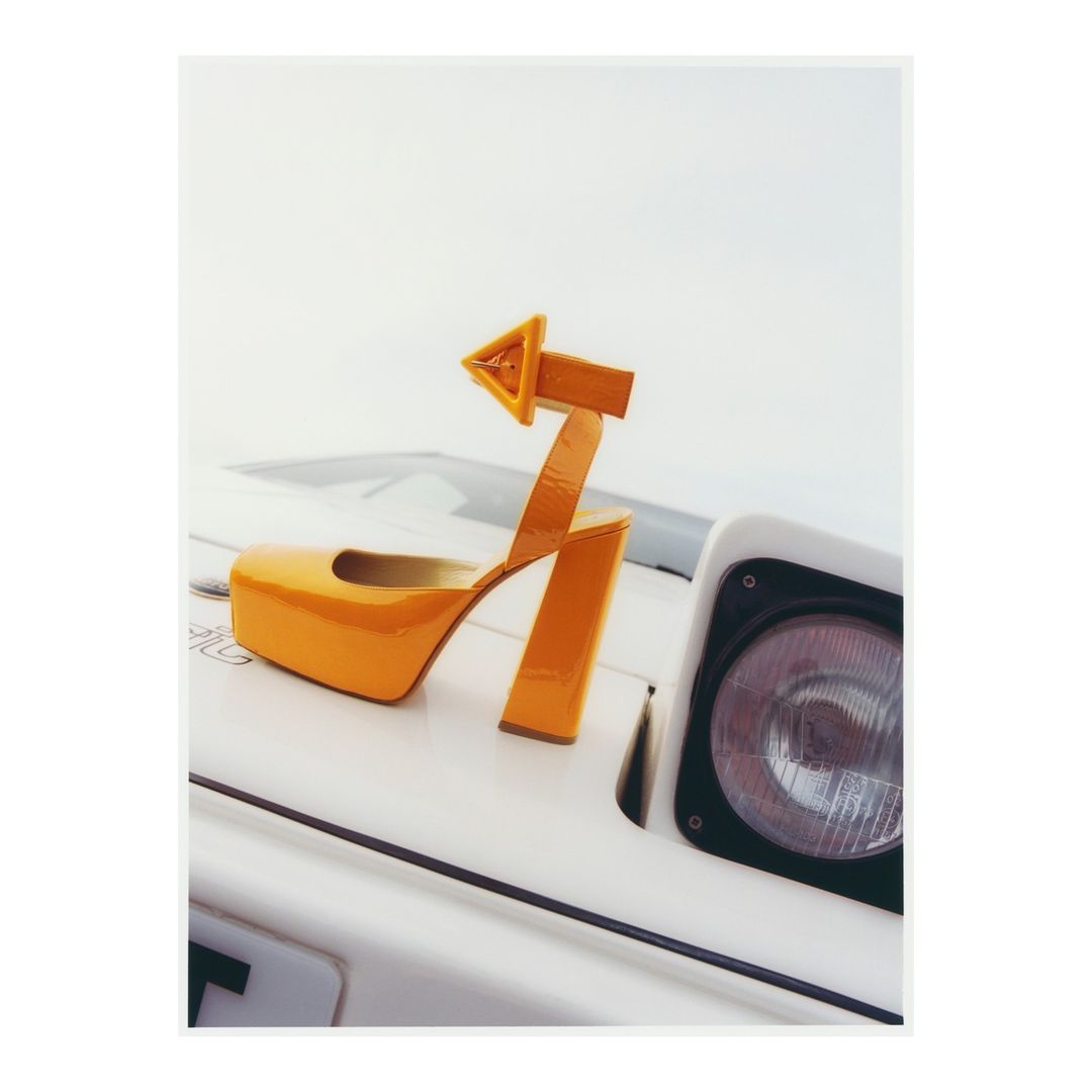 Elevated view of a single orange, patent-leather platform heel sitting on the hood of a white car against a bright, overcast sky.