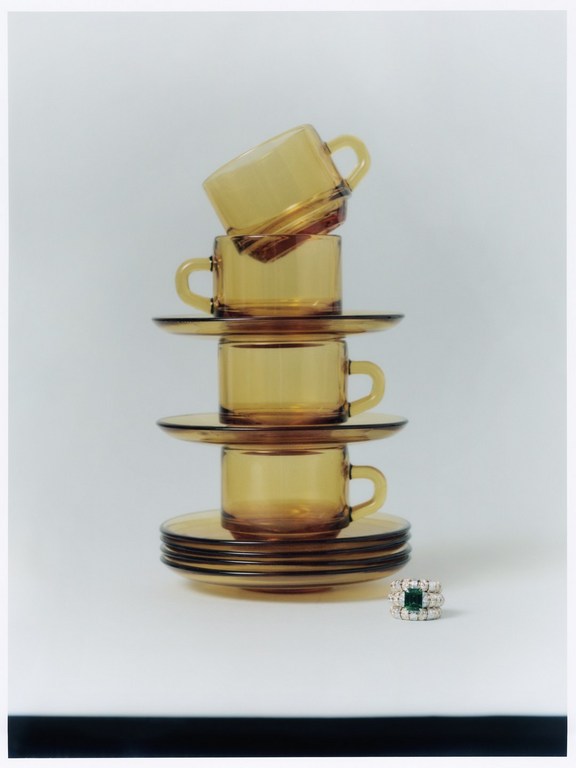Stacked amber glass tea cups and saucers form a tower next to a gemstone ring on a solid white surface, creating an elegant display.