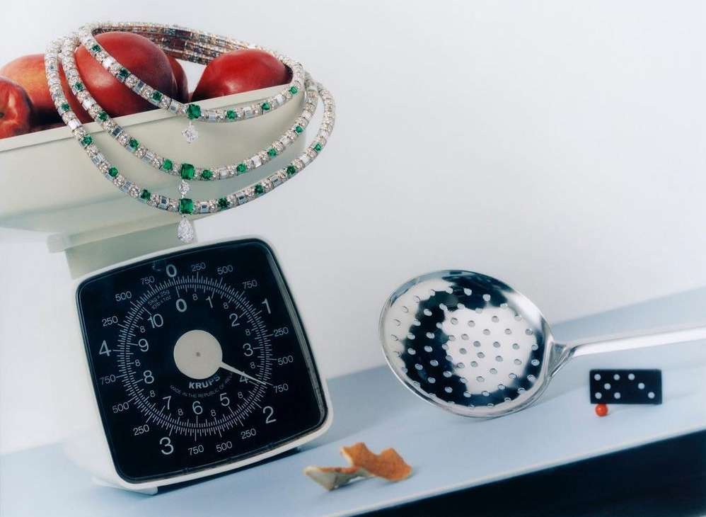 Elegant emerald and diamond necklace displayed on a white kitchen scale amidst peaches and a slotted spoon, showcasing luxury jewelry.