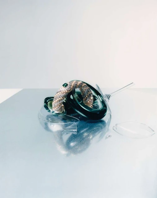 Gold woven bracelet with green gemstone on top of a smashed wine glass, sitting on a reflective surface. Modern jewelry still life.