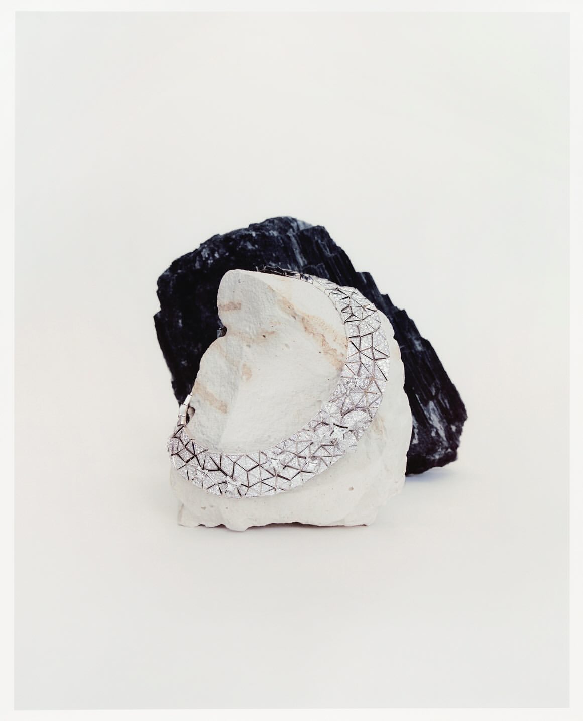 Silver bracelet with geometric pattern resting on a white rock, with a black rock behind against a plain white background. Minimalist jewelry.