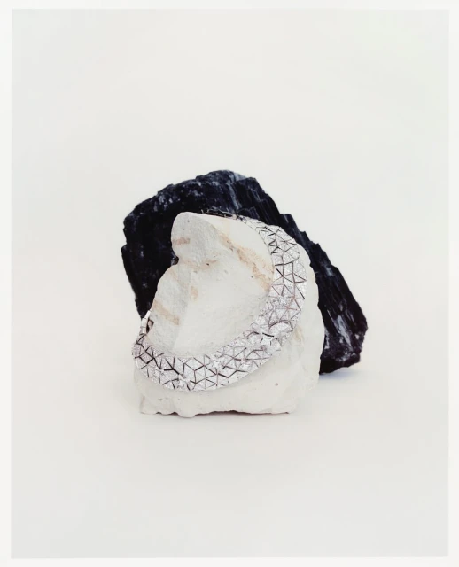 Silver bracelet with geometric pattern resting on a white rock, with a black rock behind against a plain white background. Minimalist jewelry.