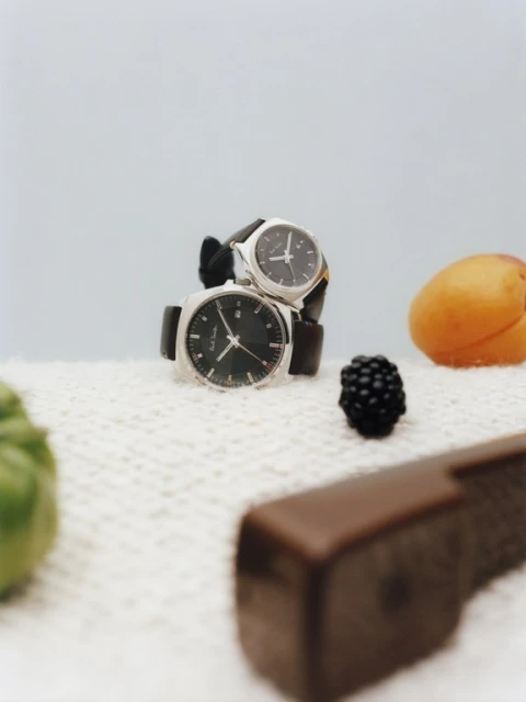 Two wristwatches are stacked on a textured white surface with fresh fruit and a chocolate bar on a light blue background.