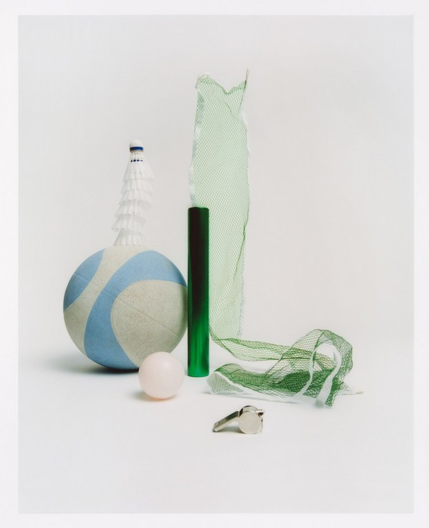 Still life photograph features a volleyball, badminton birdie, net, whistle and rubber ball on a seamless white backdrop.
