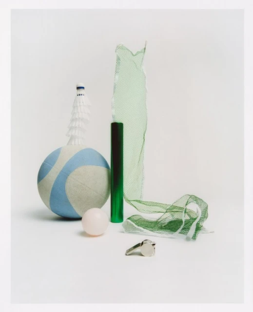 Still life photograph features a volleyball, badminton birdie, net, whistle and rubber ball on a seamless white backdrop.