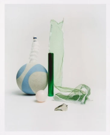 Still life photograph features a volleyball, badminton birdie, net, whistle and rubber ball on a seamless white backdrop.