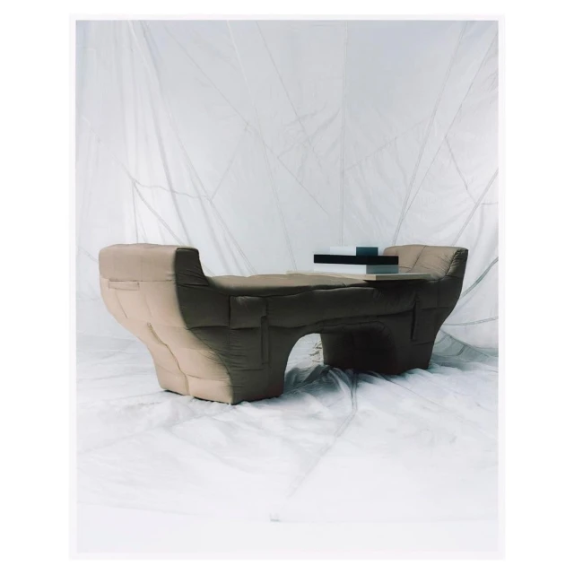 Studio shot of a modern puffy beige bench seat with a floating table element on top, set against a wrinkled white fabric backdrop.
