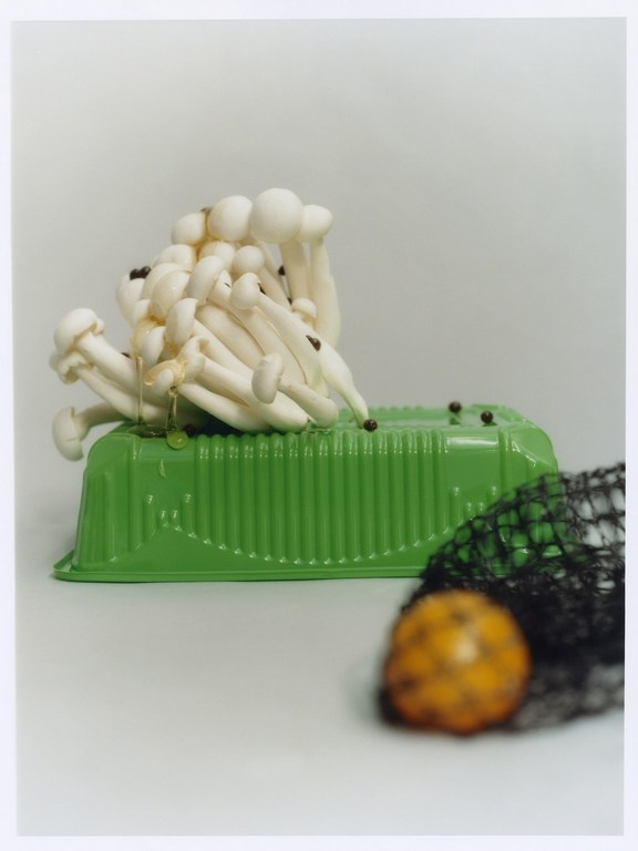 Still life photograph of a cluster of white beech mushrooms spilling from a green container onto a black net, set against a white backdrop.