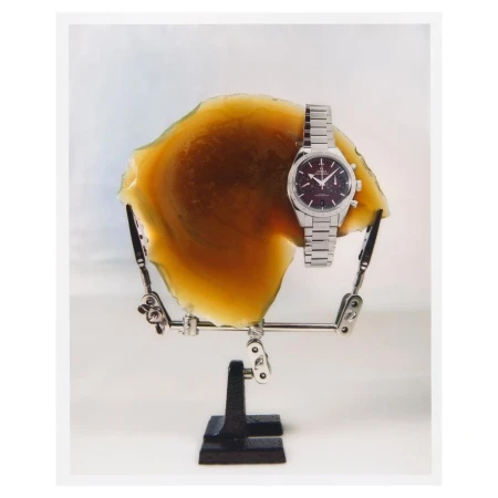 Studio product shot of a luxury silver wristwatch displayed on an adjustable jewelry stand with a slice of petrified wood as backdrop.