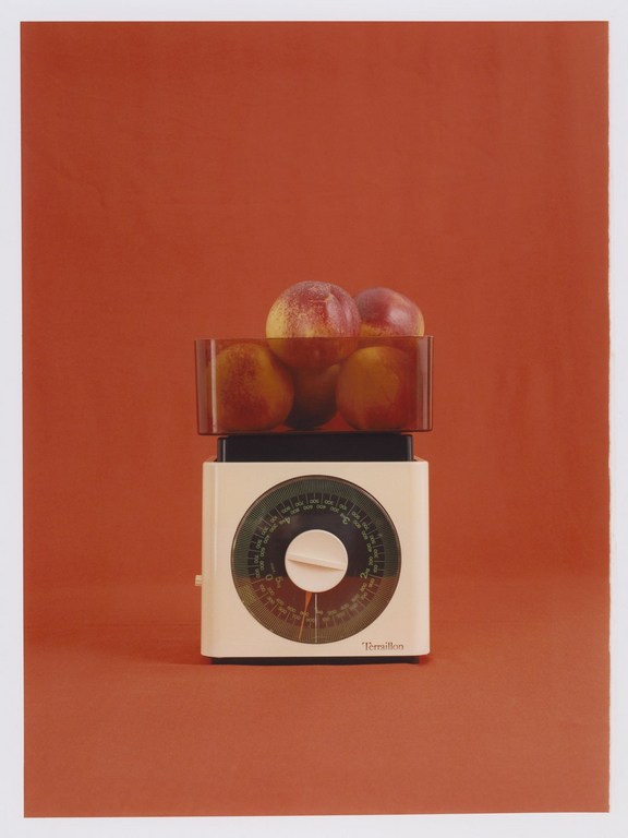 Studio shot of a vintage scale with peaches on a solid red backdrop, highlighting the scale's design and the peaches' vibrant colors.