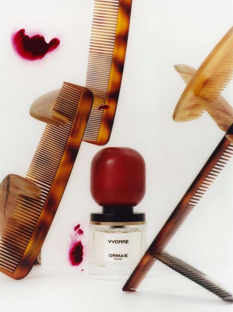 Still life shot of Yvonne ORMAIE Paris perfume with amber combs on a white backdrop with red liquid splashes creating visual interest.