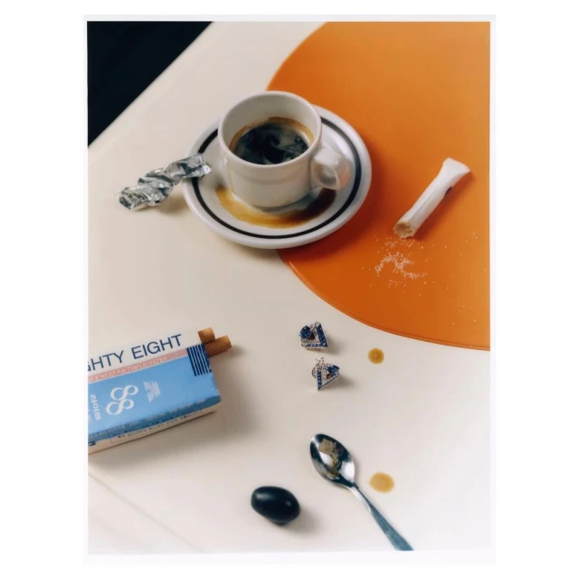 A cup of black coffee on a saucer with a cigarette pack, sugar, and a spoon on a white table with an orange circle.