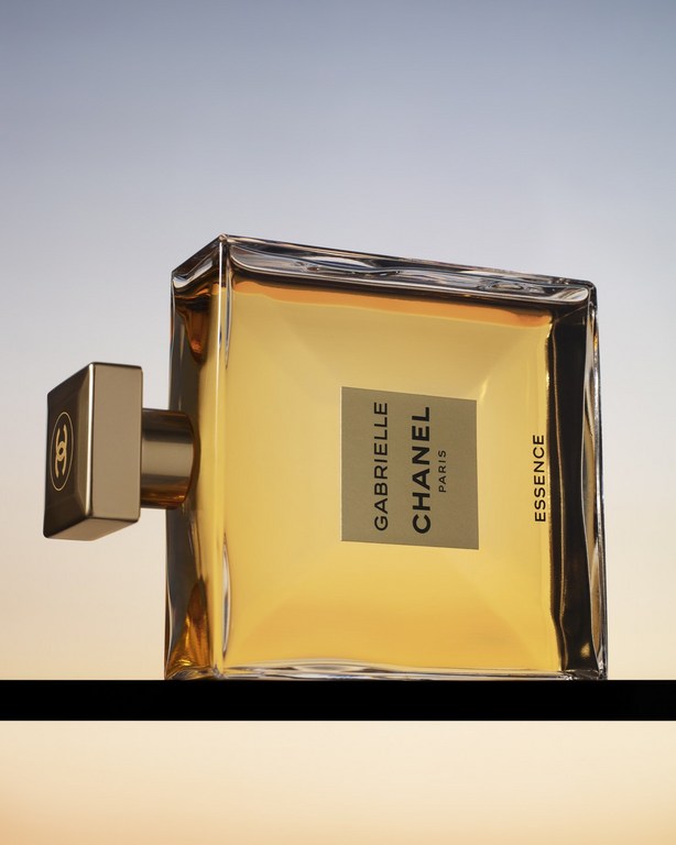 Close-up shot of Chanel Gabrielle Essence perfume bottle with golden liquid on a black surface against a gradient background.