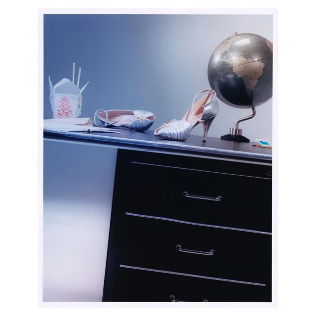 Fashion still life on a black dresser with a pair of silver high heel shoes, a takeout box, and a globe in an indoor setting.