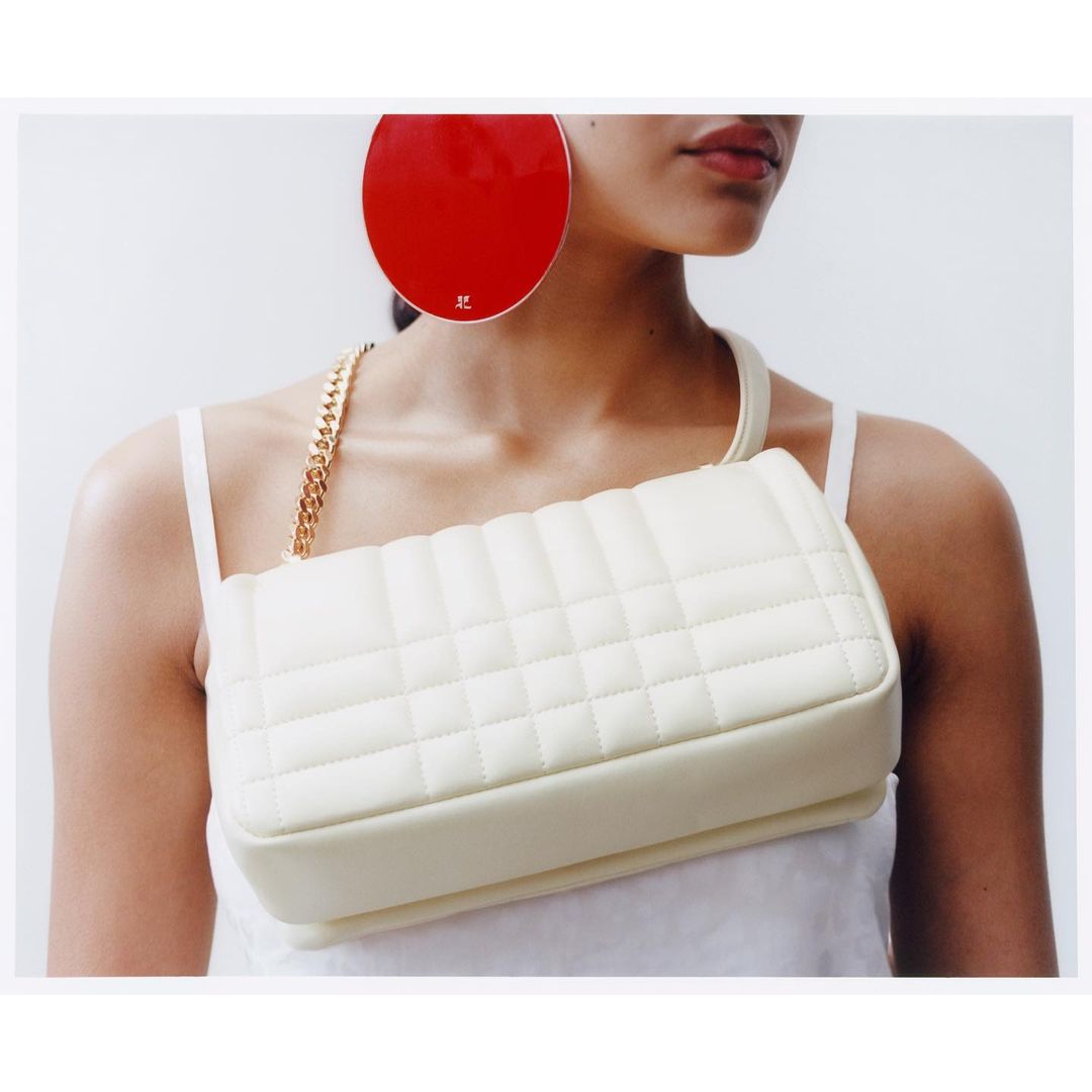 Fashion shot featuring a model wearing large red disc earrings, a white top, and a quilted off-white shoulder bag with a gold chain.