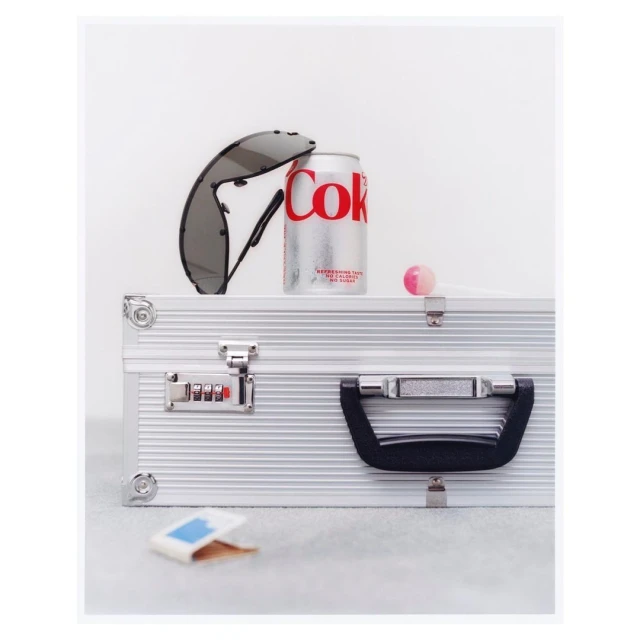 Still life featuring a can of Coke, sunglasses, a pink lollipop, and a small package, all arranged on top of a silver briefcase.