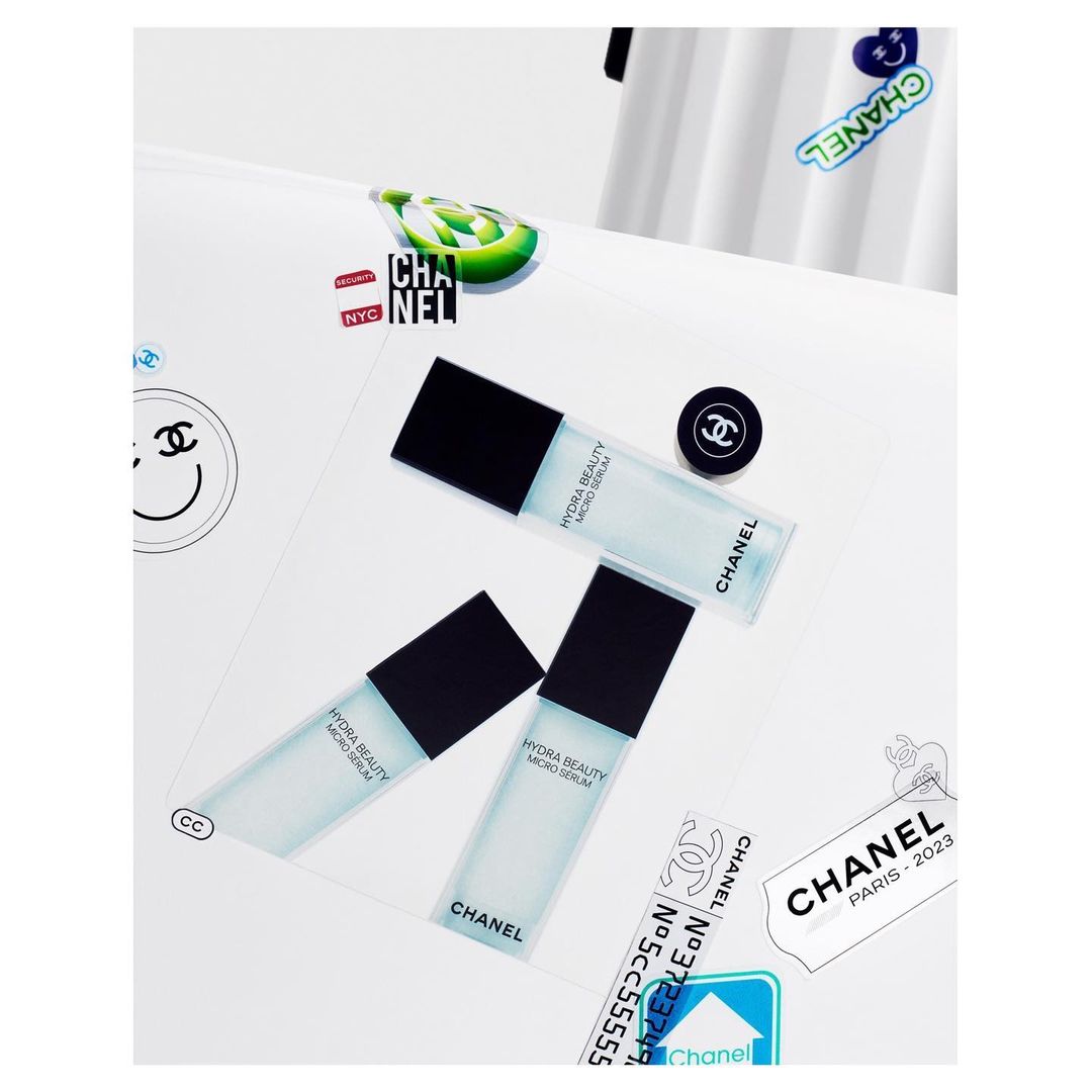 Three bottles of Chanel Hydra Beauty Micro Serum are arranged on a white surface with Chanel-themed stickers and a button, showcased in a studio shot.