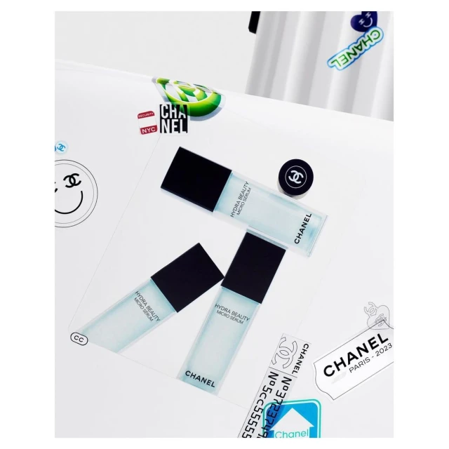 Three bottles of Chanel Hydra Beauty Micro Serum are arranged on a white surface with Chanel-themed stickers and a button, showcased in a studio shot.