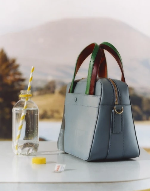 Blue satchel handbag on a white table with a bottle of water and a straw, set against a blurred background of mountains and a lake.