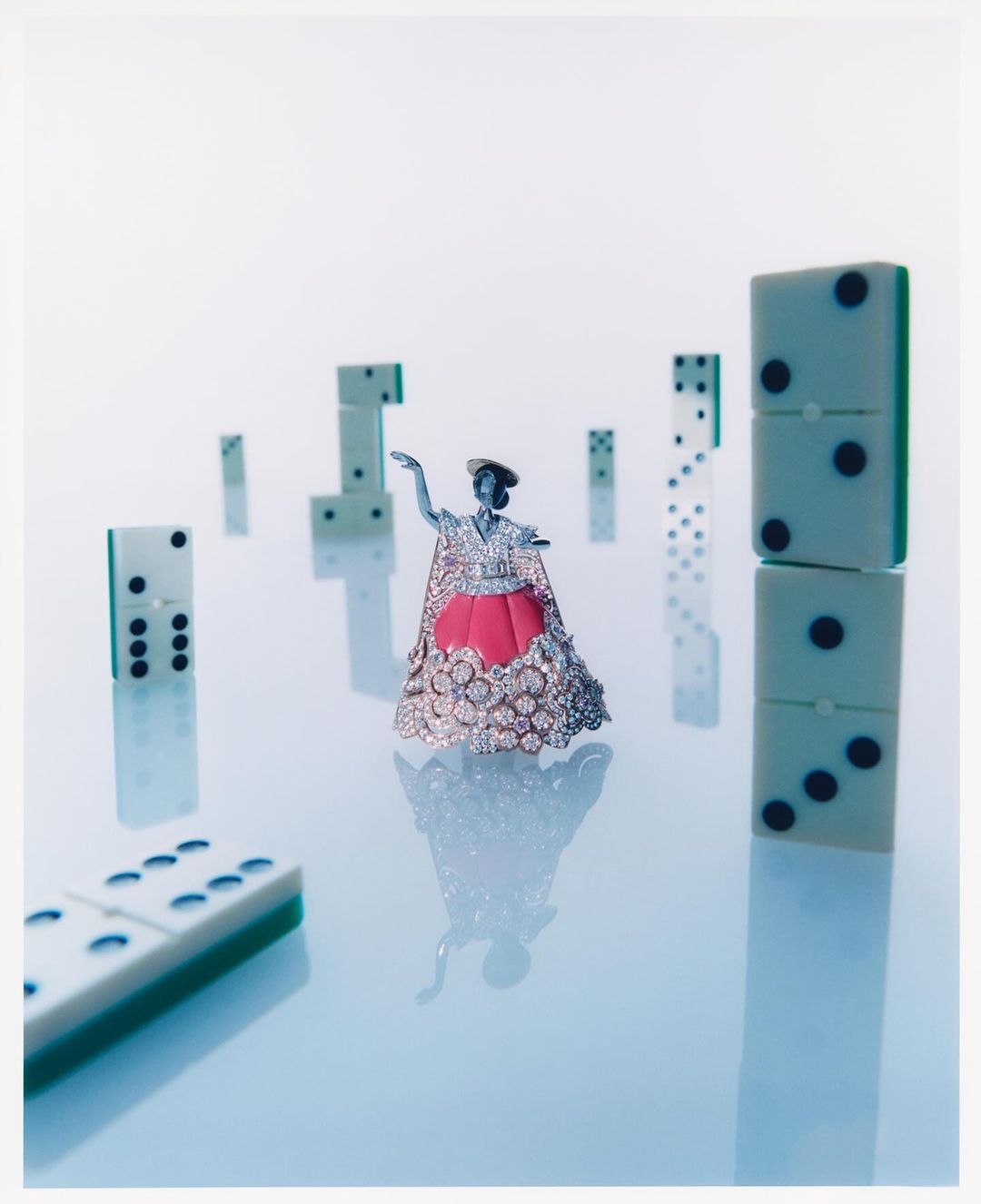 Surreal still life with a tiny female figure in a cape among dominoes, set against a gradient blue and white backdrop, creating an otherworldly scene.