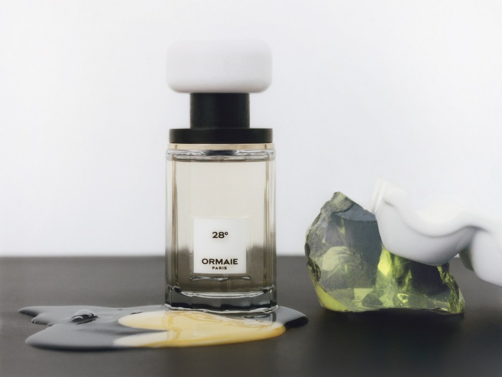 A bottle of Ormaie Paris perfume with a white cap and green stone on a black surface against a bright background.