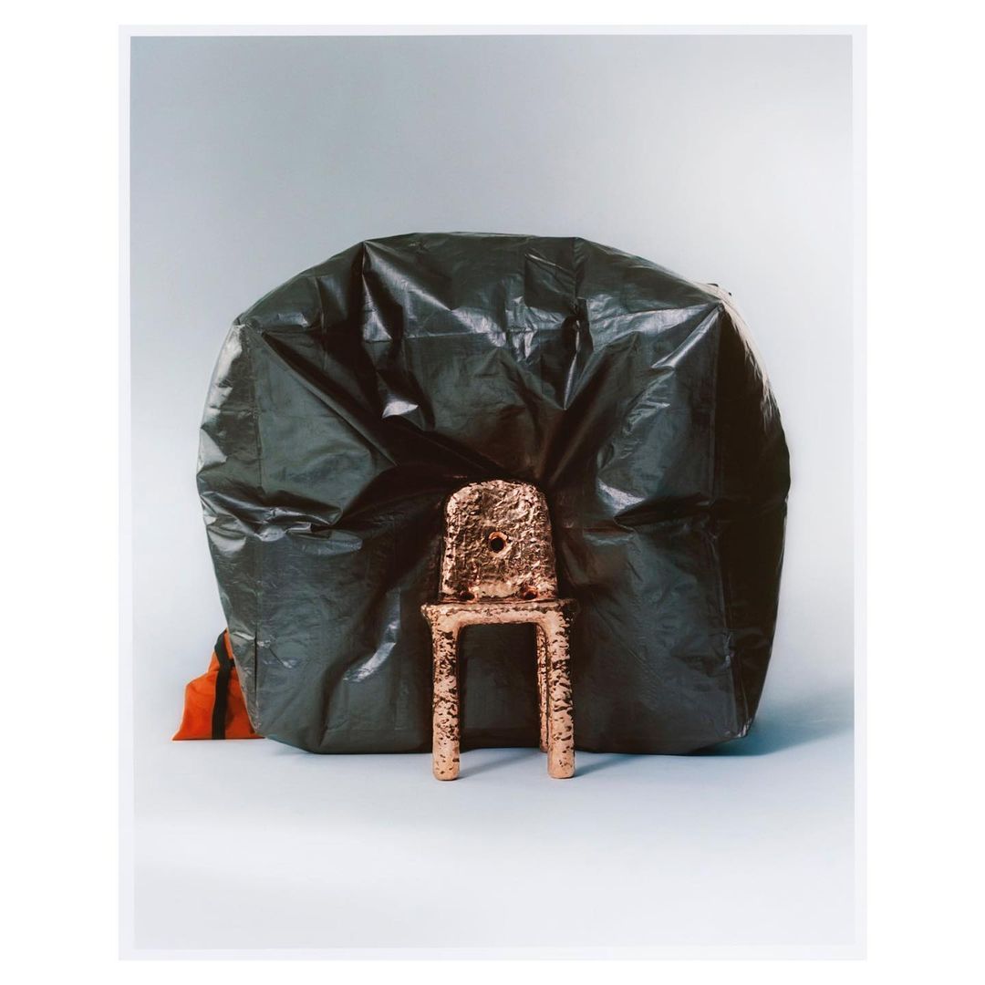 Still life of a miniature, textured, gold chair placed in front of a large black plastic bag against a grey-white background.