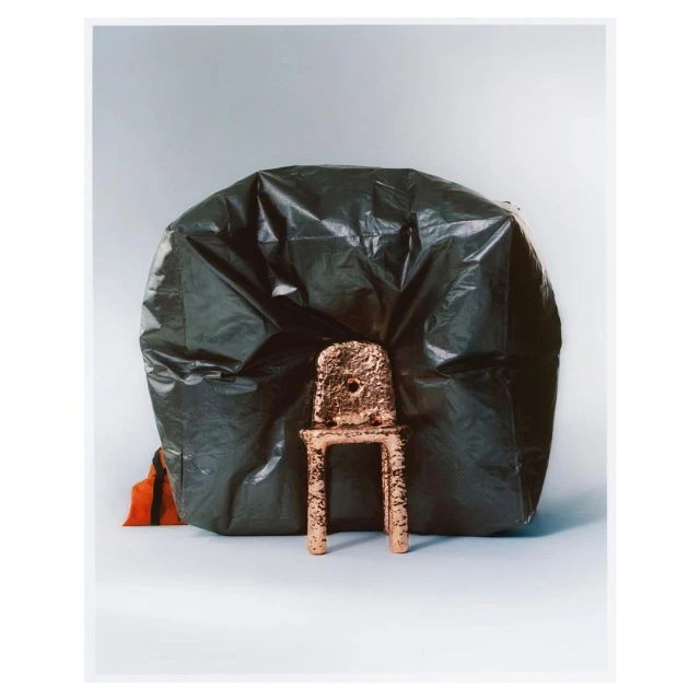 Still life of a miniature, textured, gold chair placed in front of a large black plastic bag against a grey-white background.