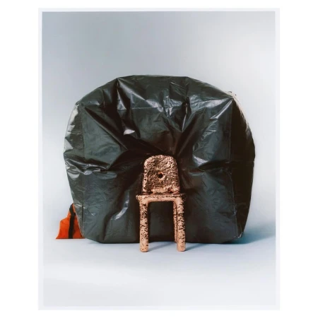 Still life of a miniature, textured, gold chair placed in front of a large black plastic bag against a grey-white background.