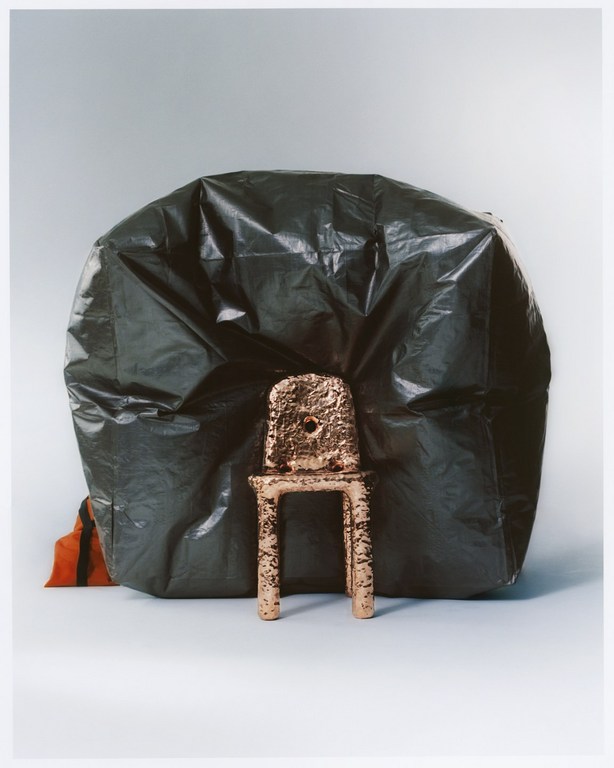 Abstract studio still life photography: a copper-colored chair is placed in front of a large, dark green plastic garbage bag.
