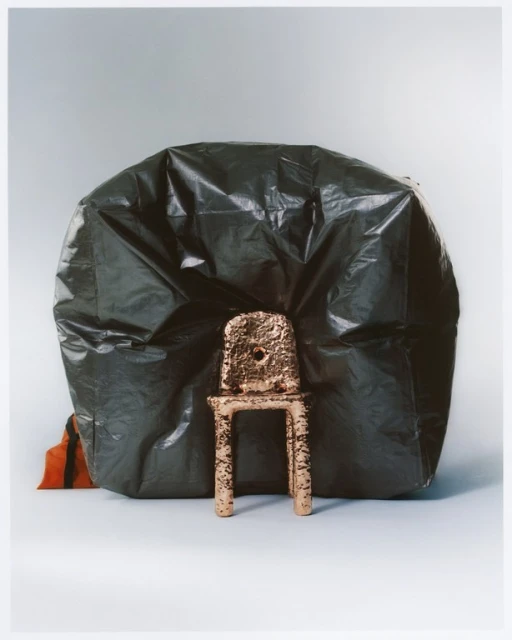 Abstract studio still life photography: a copper-colored chair is placed in front of a large, dark green plastic garbage bag.