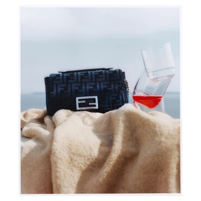 Fendi handbag with logo pattern on sand with drink. Designer purse on beige towel near transparent glass of orange liquid.