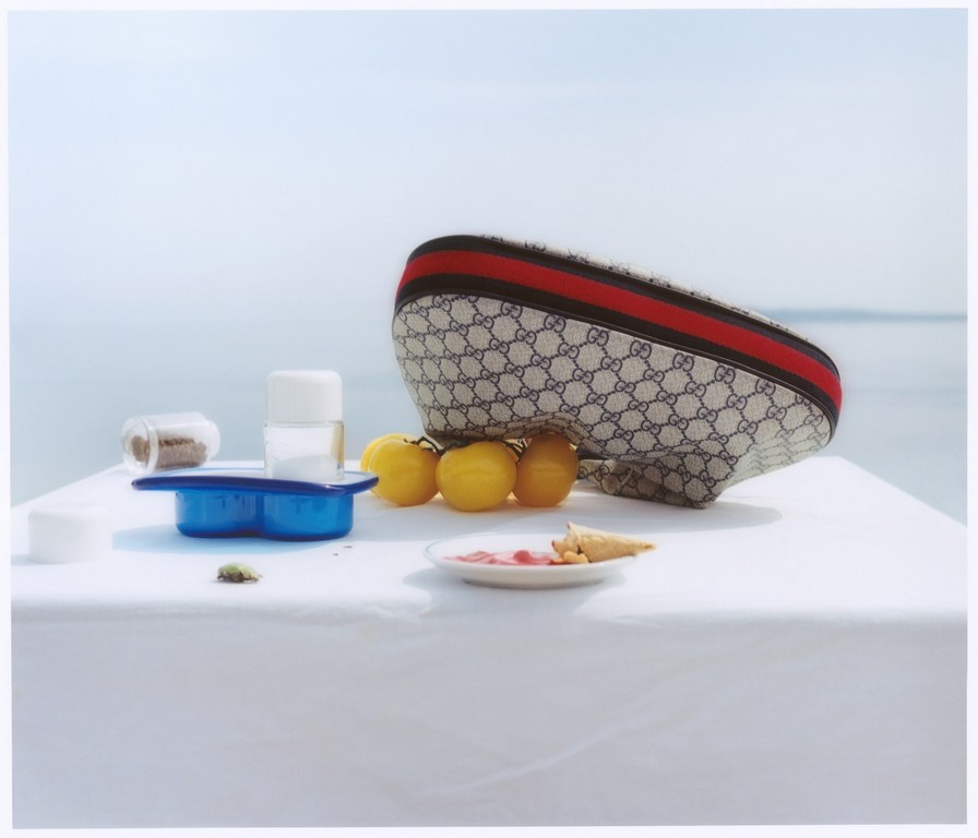 Still life on a white table against a light blue background featuring a Gucci bag, lemons, candies, containers, and ocean view.
