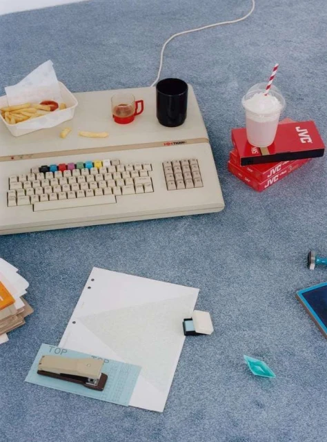 Retro setup: vintage computer with colorful keyboard, fries, drink, office supplies on a grey carpet. Nostalgic tech and modern elements.