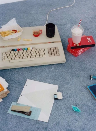 Retro setup: vintage computer with colorful keyboard, fries, drink, office supplies on a grey carpet. Nostalgic tech and modern elements.
